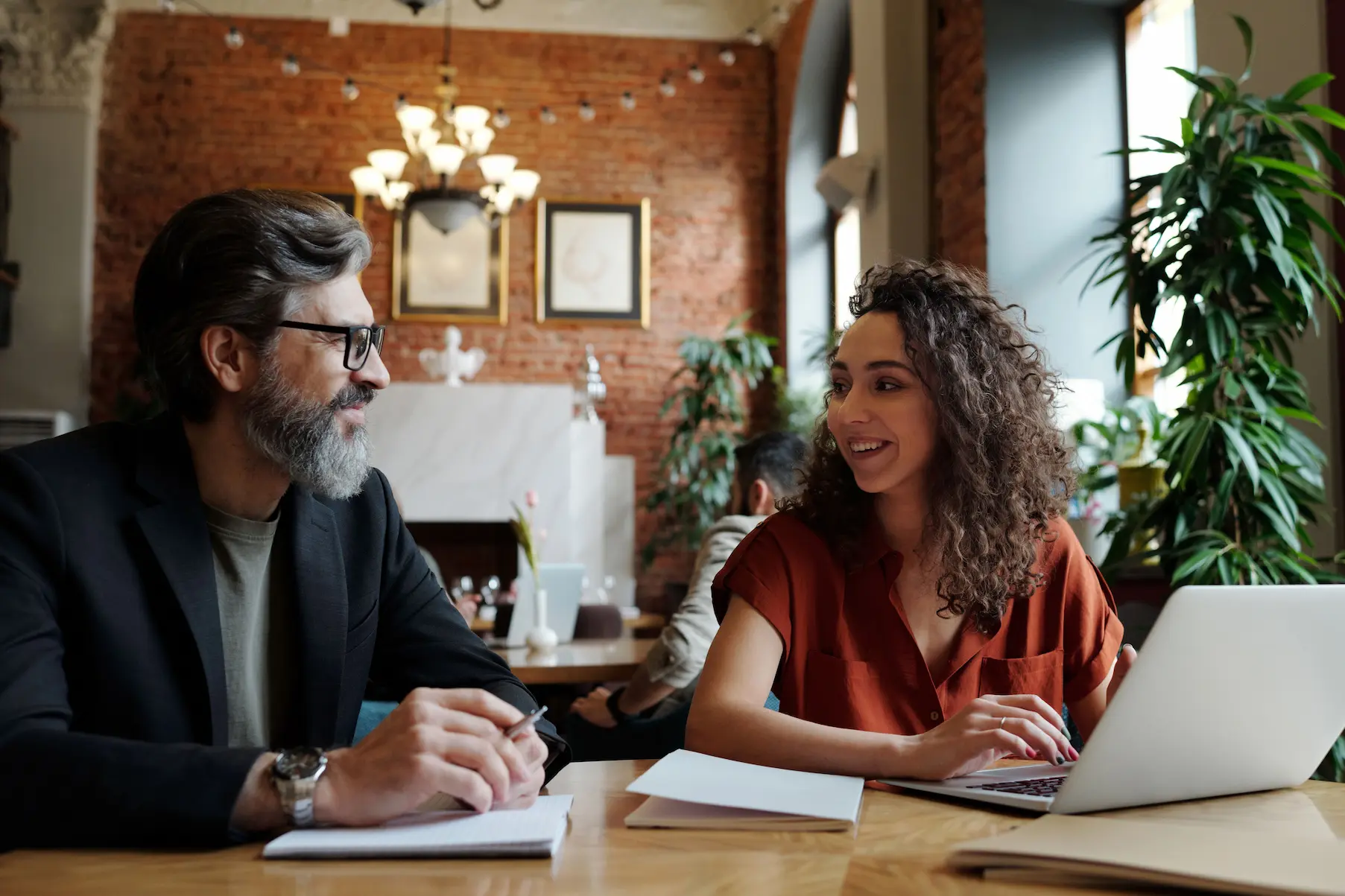 Two people engaged in a discussion at a table, with a laptop and notebooks, in a cozy, plant-filled cafe setting.