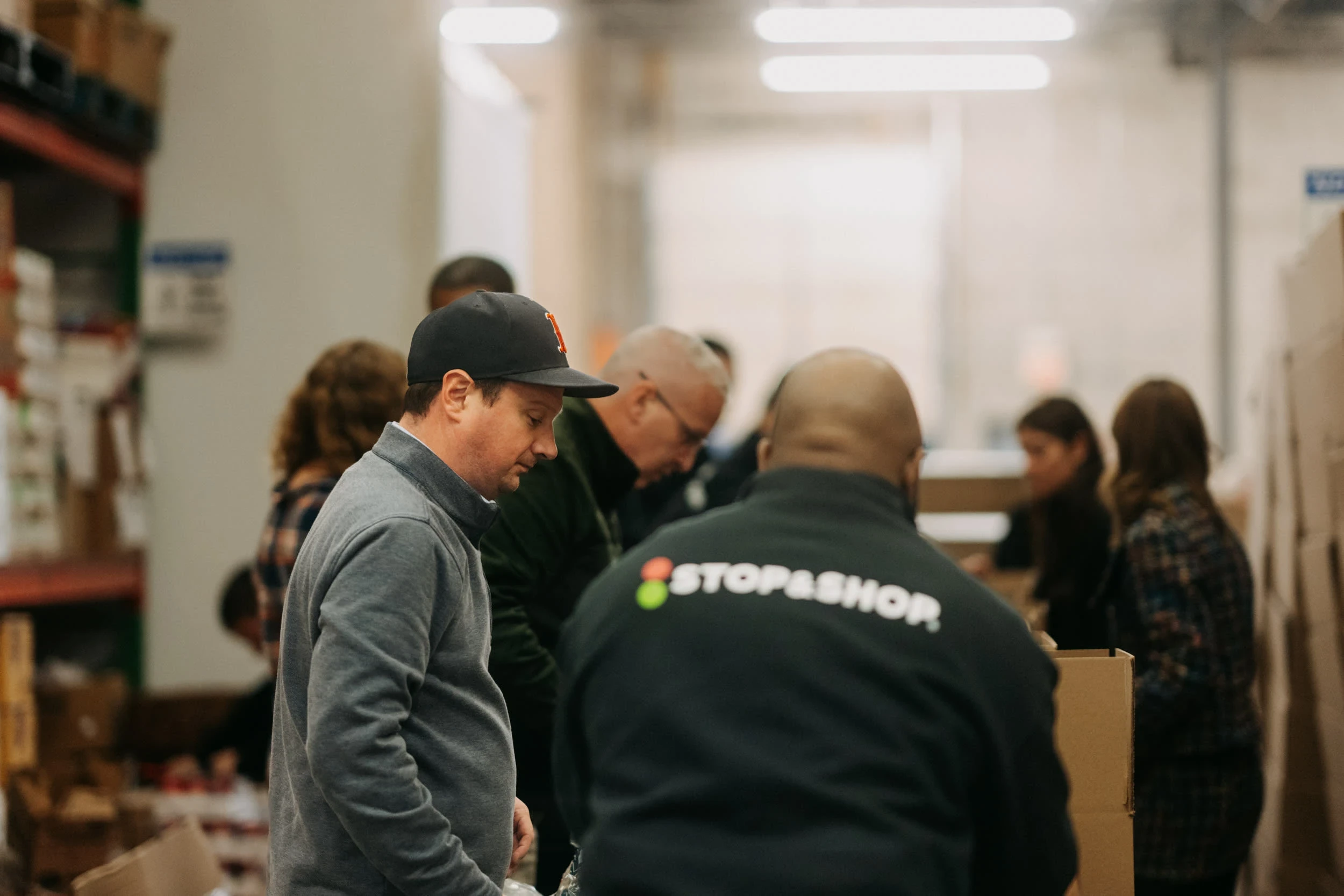 A candid photograph of a group of people inside a warehouse or distribution center. In the foreground on the left, a man in a gray sweatshirt and a dark baseball cap is looking down and focusing on something. In the center, the back of a person wearing a dark jacket with the white and green Stop & Shop logo is visible. Other people are gathered around, and the background features brightly lit, tall metal shelving with stacked boxes.