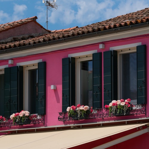 A pink house with three upper-floor windows, green shutters, and flower boxes filled with pink and white flowers under a tiled roof.