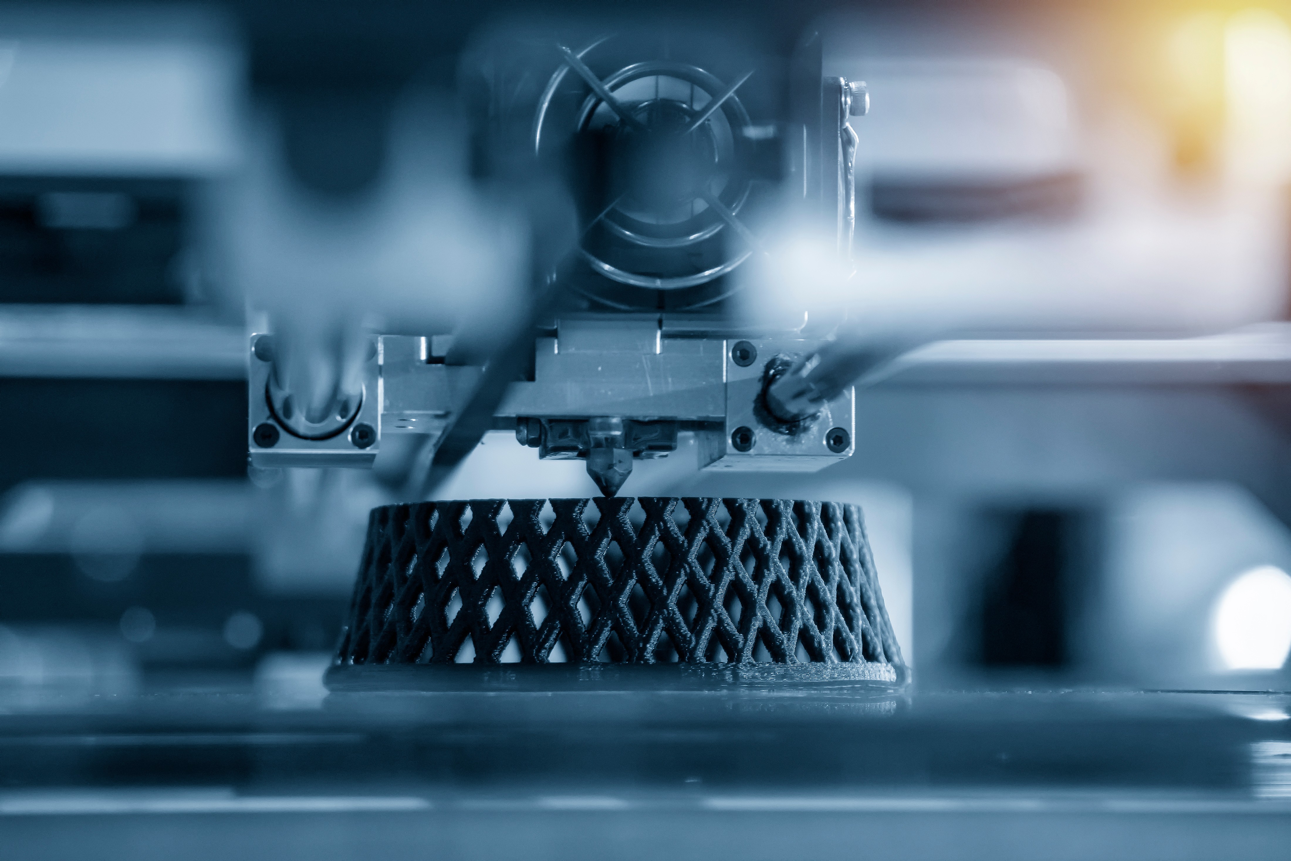 Close-up of a 3D printer nozzle printing a black lattice-structured component, in blue tones