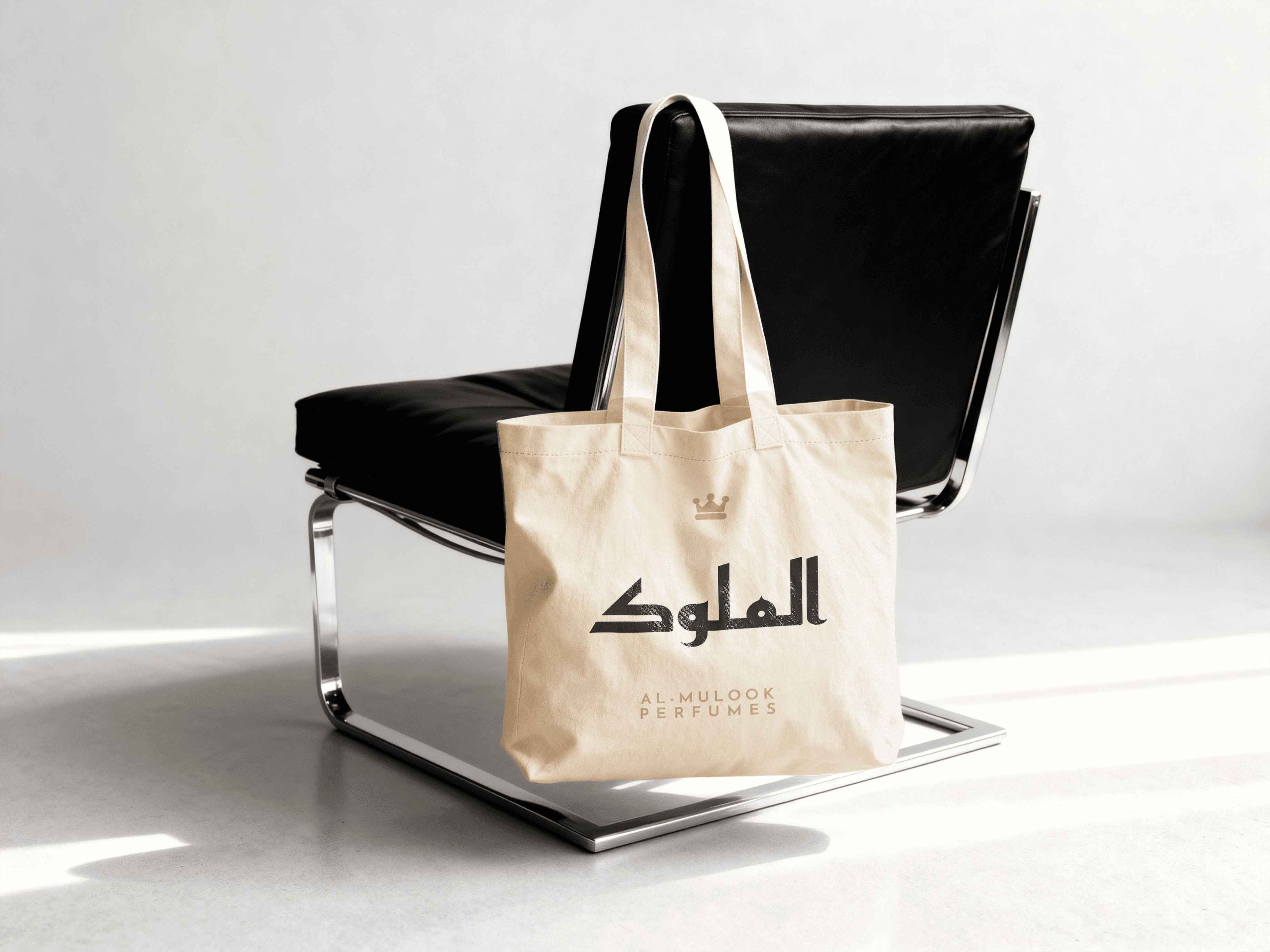 Beige Al-Mulook branded tote bag with Arabic logo and “Smell Like Royalty” tagline placed on a modern black chair in a studio setting.
