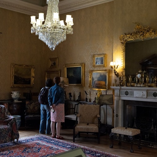 Two people in a vintage room with antique furniture, paintings on the walls, a large chandelier, and an ornate fireplace.