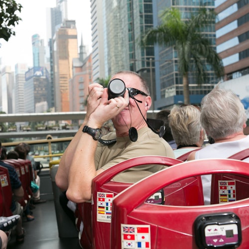 在一个充满高楼和棕榈树的城市里,一位男士与其他游客一起乘坐开放式观光巴士时拍摄照片。
