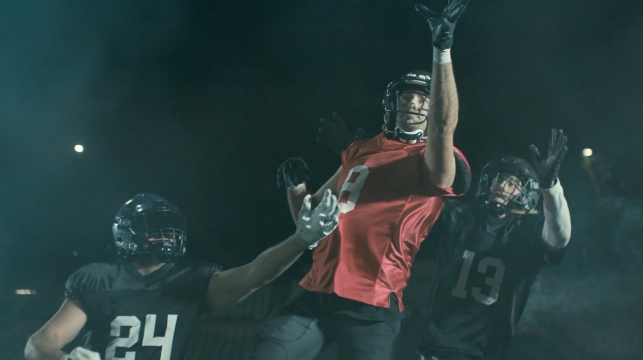 Three football players under stadium lights: a receiver in red leaping between two defenders in black.