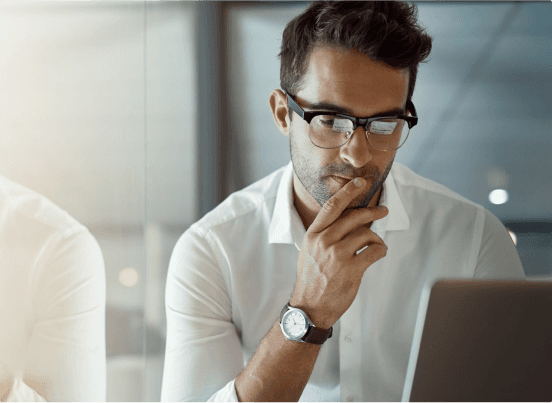 Homem utilizando óculos e camisa branca, olhando atento para o computador, pensando
