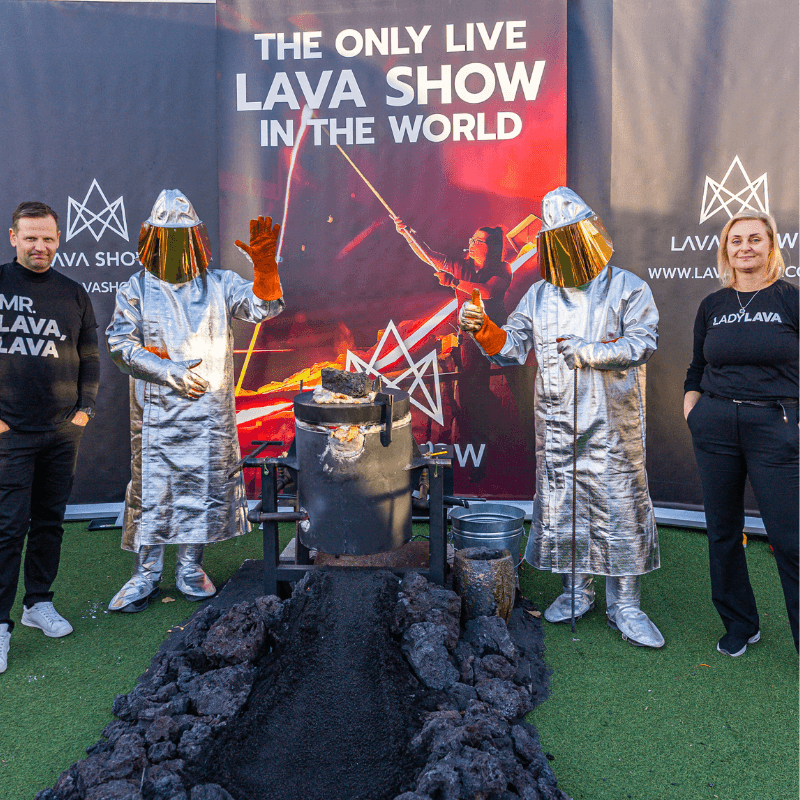 Group photo at the Taste of Iceland event in Toronto. On the left is Lava Show founder, Júlíus Ingi Jónsson, AKA Mr. Lava Lava, two lava melters dressed in silver heat-protection suits operating a mobile lava furnace, and on the far left is Lava Show founder Ragnhildur Ágústsdóttir, Lady Lava Lava. Cooled lava rocks are arranged in front of the furnace, with Lava Show banners in the background.