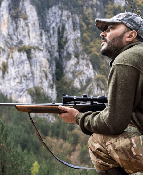 A man in a camouflage outfit holds a rifle while sitting against a rocky backdrop in a lush green landscape.