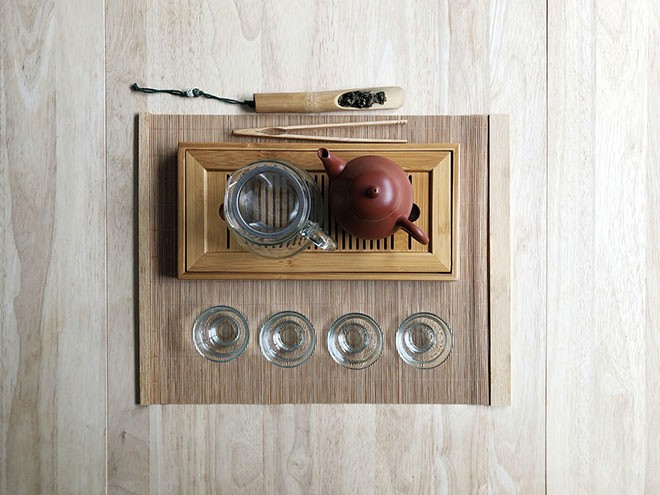 A wooden tea tray with a reddish-brown teapot and four empty clear glasses rests on a light-colored wooden surface. A pair of chopsticks and a small wooden spoon with dark speckles lie beside the tray. A dark cord is visible near the top left corner. The tray is rectangular and has a light wood grain pattern.