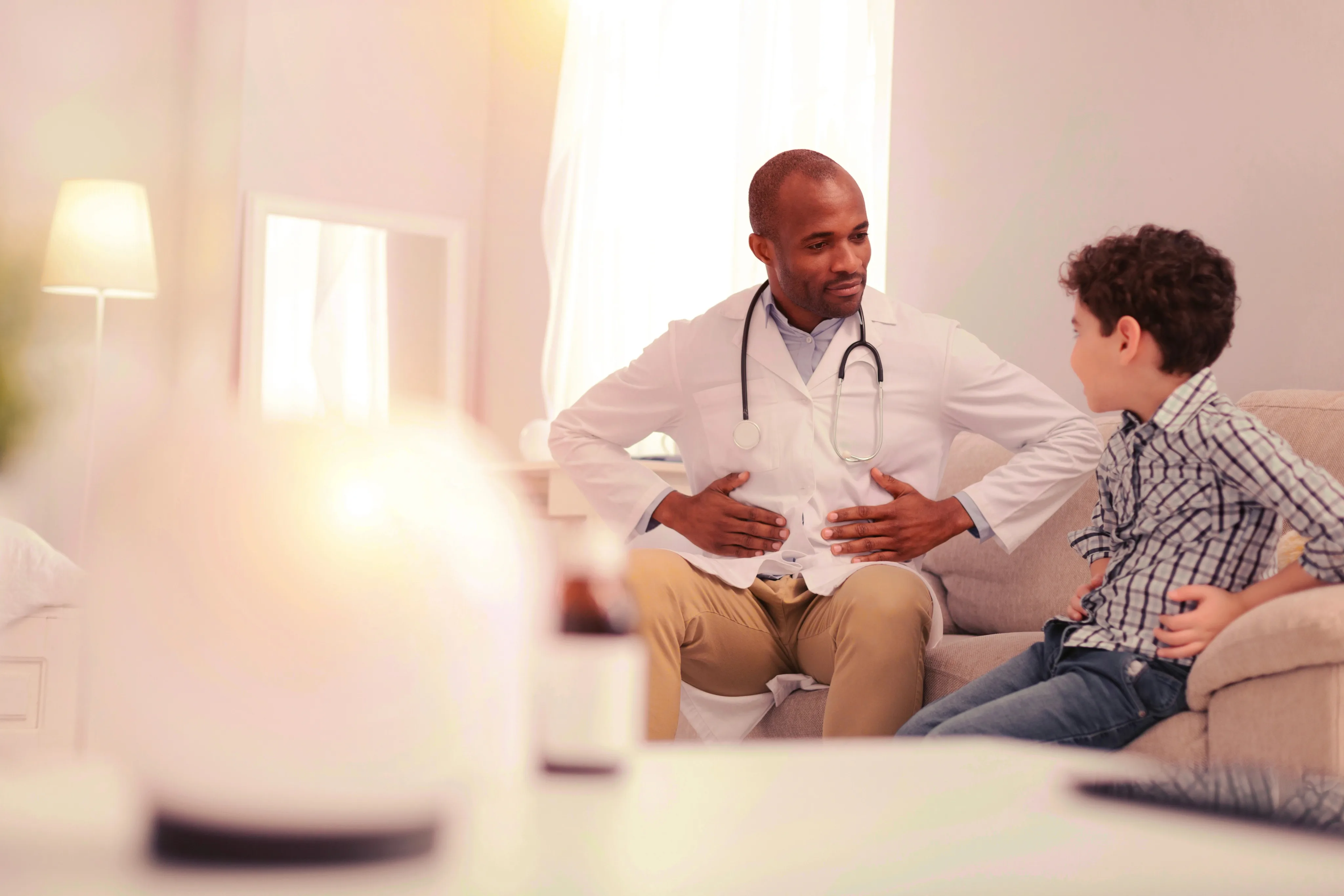 A male doctor wearing a white coat and stethoscope sits on a couch while talking with a young boy in a checkered shirt during a casual home consultation.