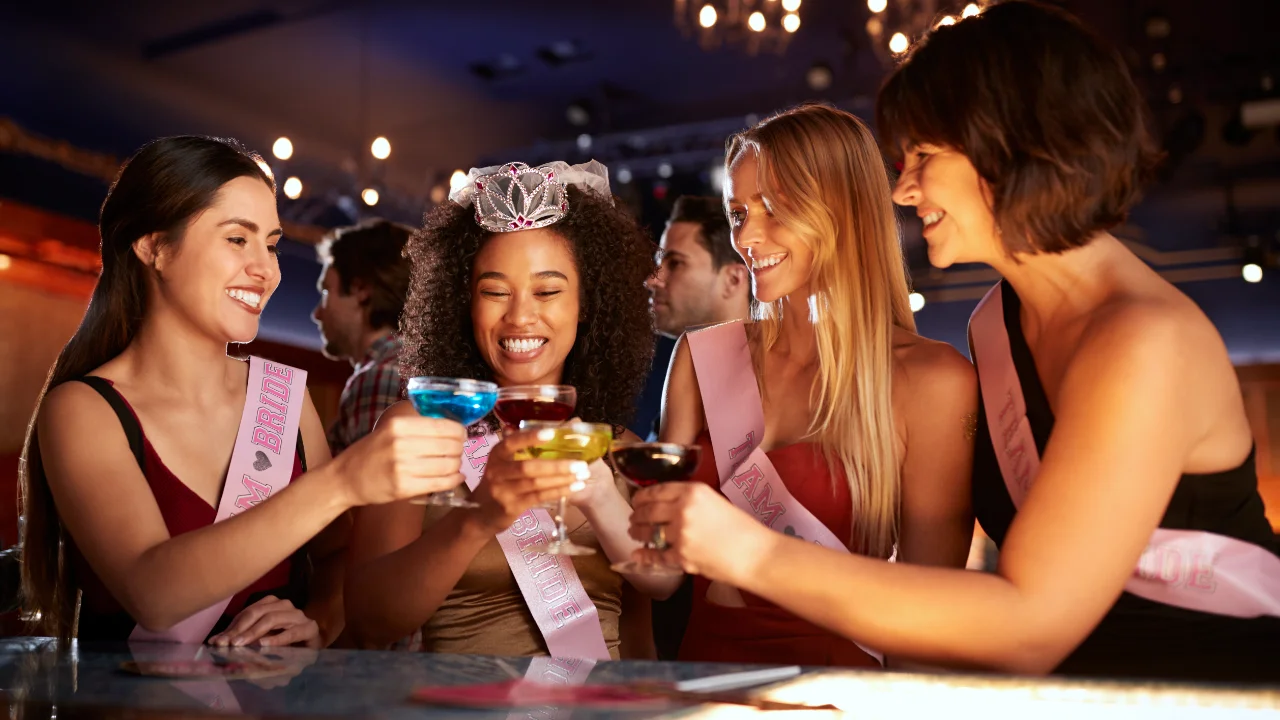 Quatre femmes souriantes, vêtues de ceintures roses "Team Bride", trinquent joyeusement avec des cocktails colorés dans.