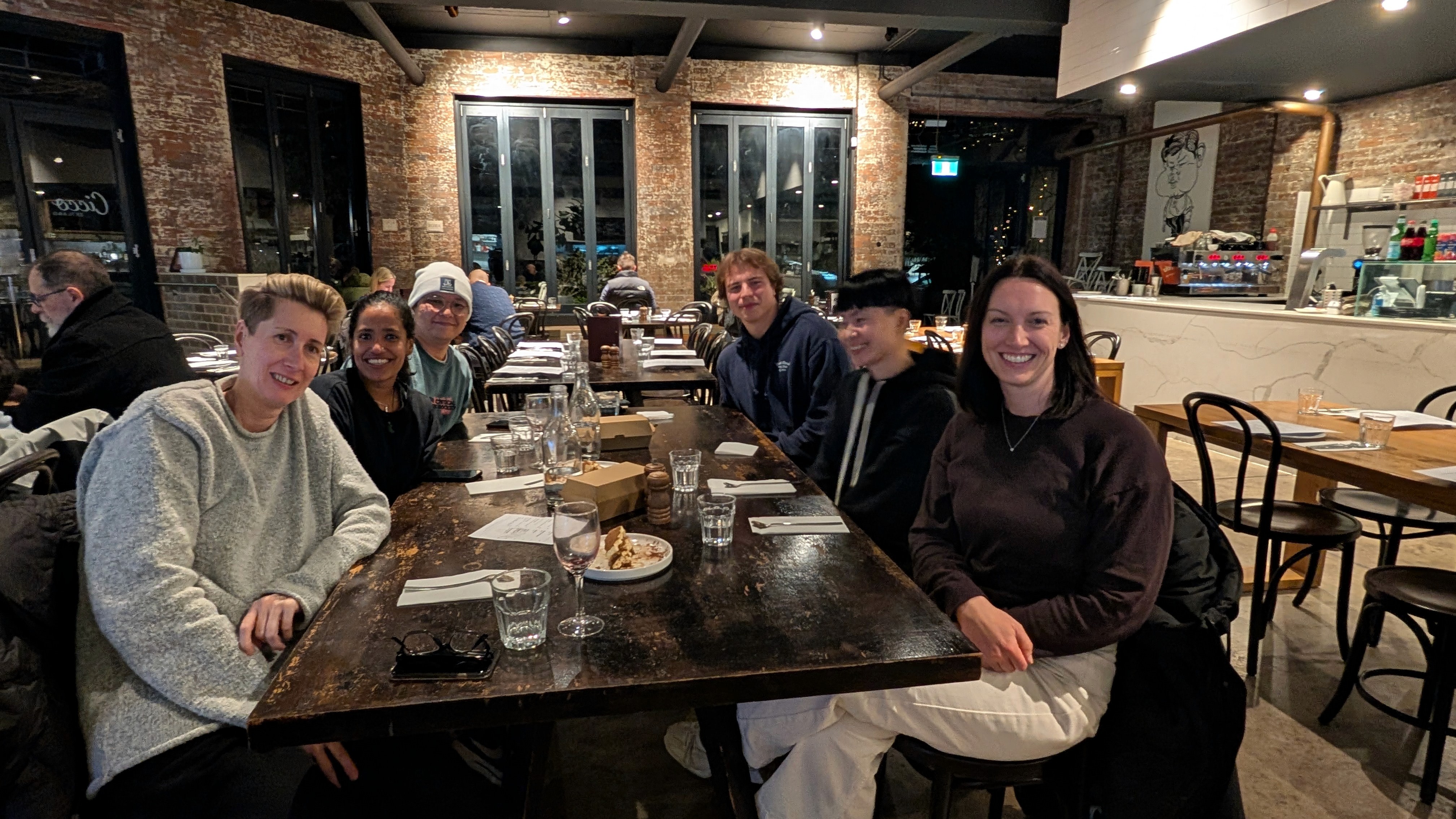 Six people seated around a long wooden table in a brick-walled restaurant at night, smiling at the camera.