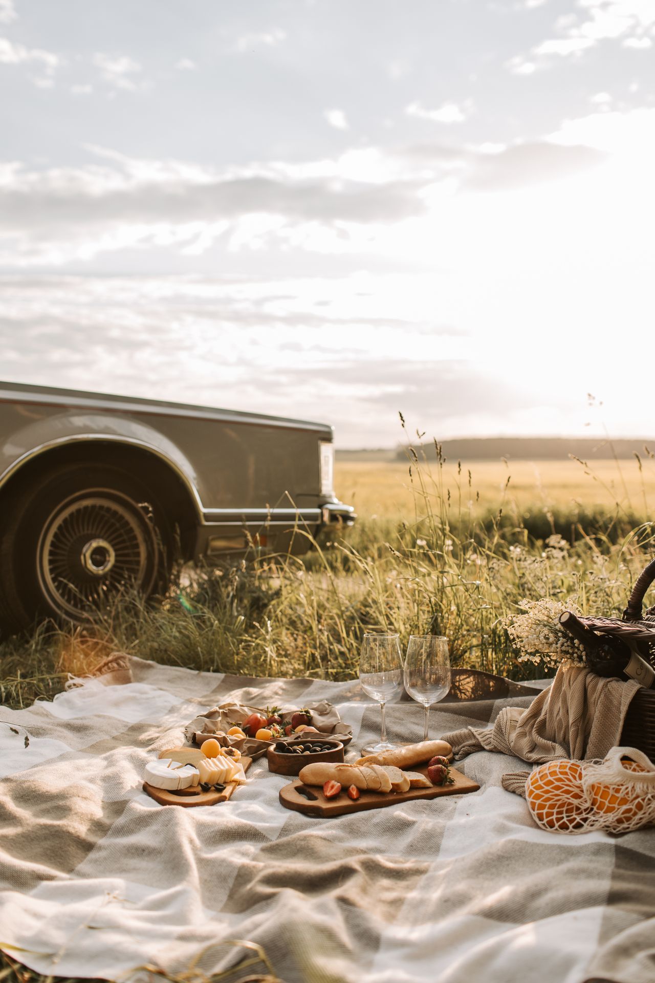 Picnic with wine, bread, and cheese spread on a blanket next to a classic car in a grassy field at sunset.