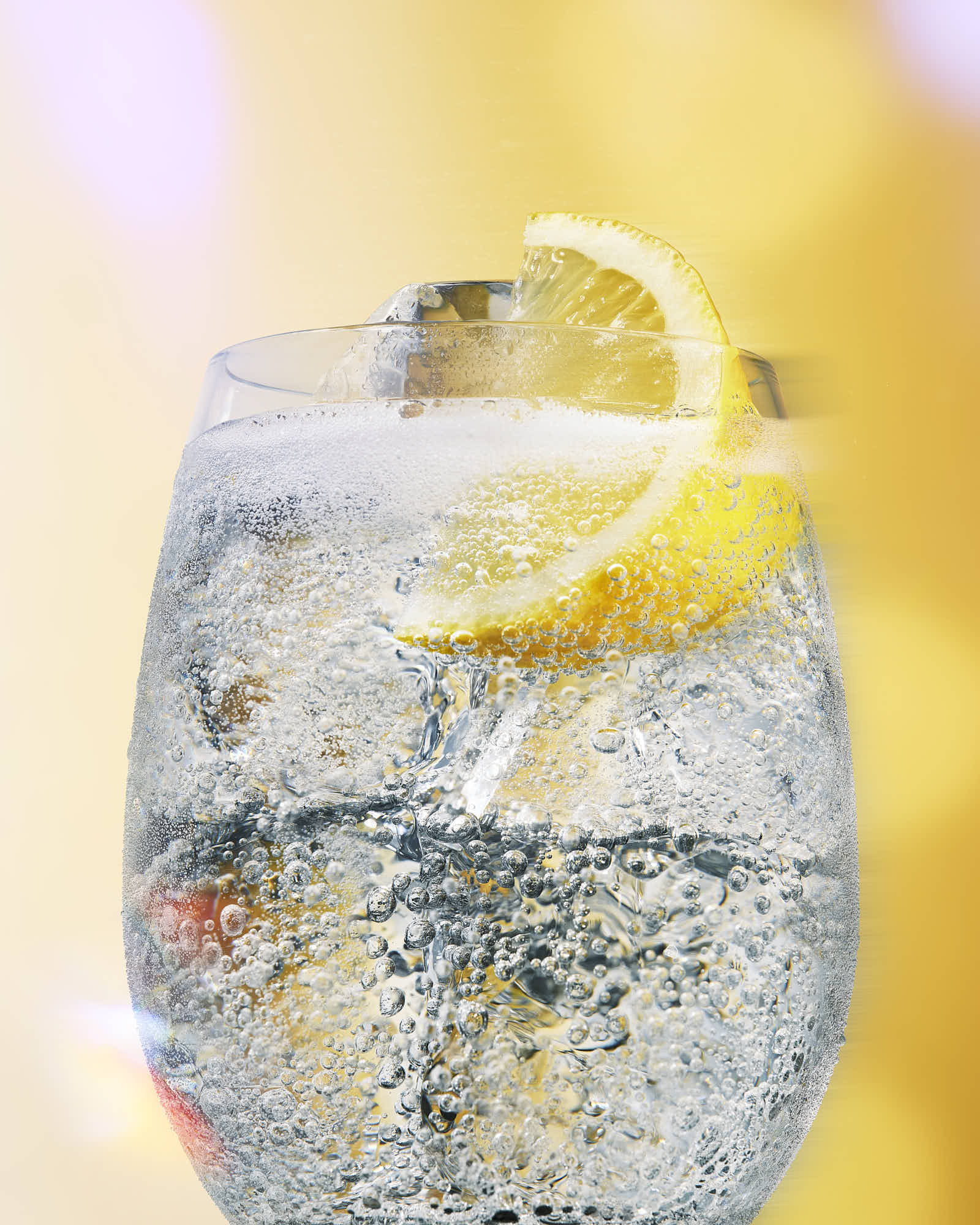 A close-up of a sparkling drink with a lemon slice, set against a soft, yellow background.