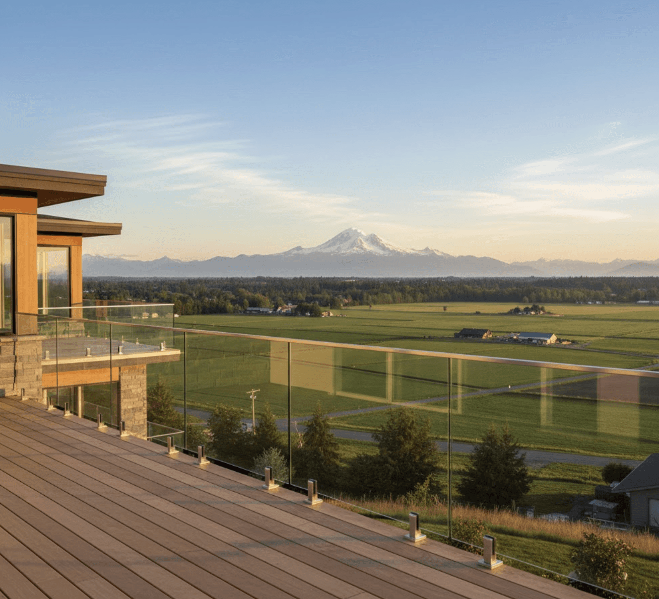 Modern frameless glass railing on a residential deck overlooking the Fraser Valley with Mount Baker visible in the distance, golden hour lighting, lush green agricultural fields in the mid-ground, contemporary Pacific Northwest home architecture, clear blue sky with wispy clouds, wooden deck flooring, clean minimal composition emphasizing the transparent glass panels and unobstructed mountain views, photorealistic style, warm natural lighting, professional architectural photography aesthetic, 16:9 aspect ratio, high clarity and depth of field, no text or overlays