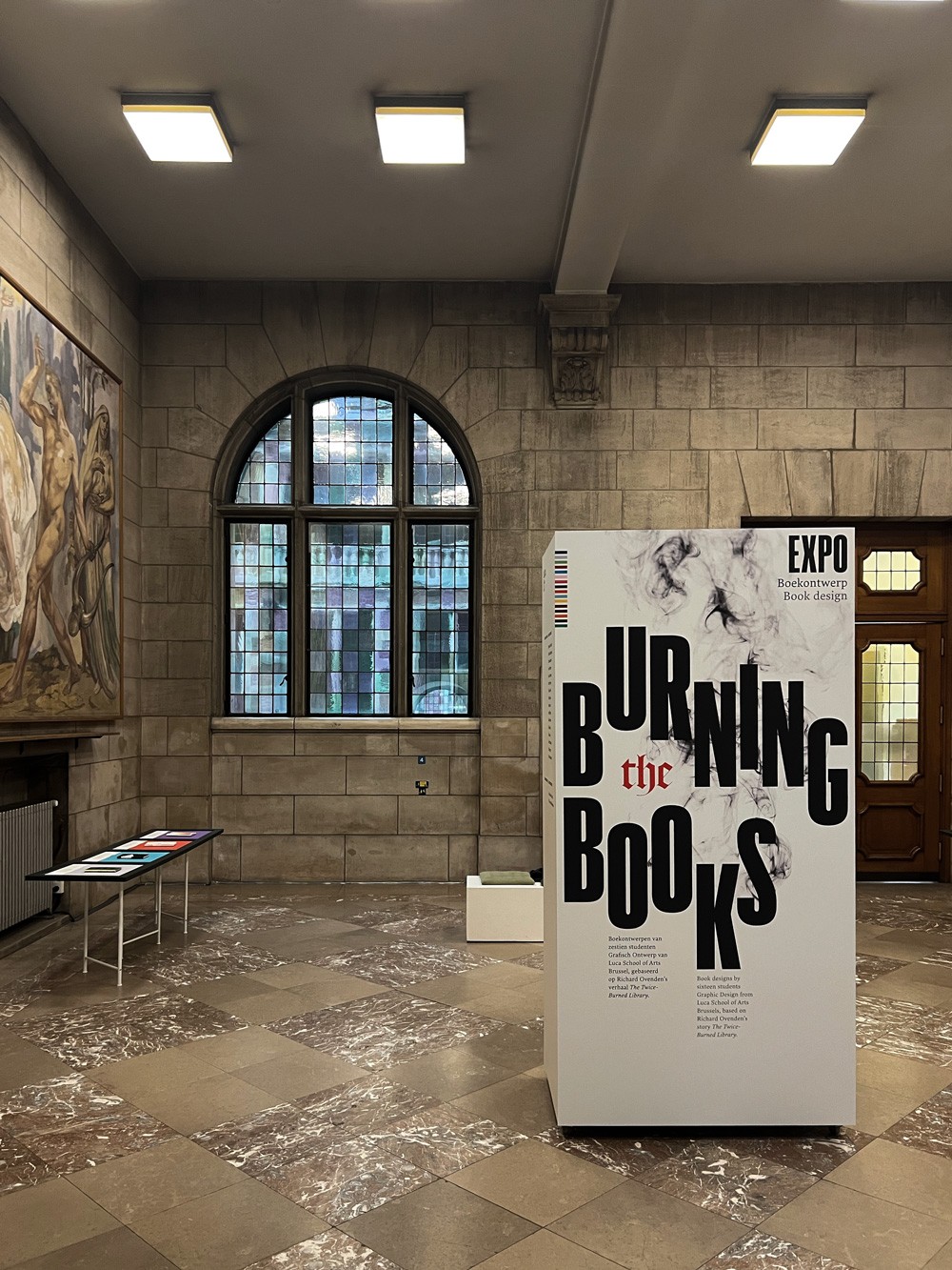 Een uitgezoomd beeld van de expositie waarop de tafels met boeken en een groot informatiebord met de titel "Burning the Books" te zien zijn. Het beeld vat de volledige context van de tentoonstelling samen.