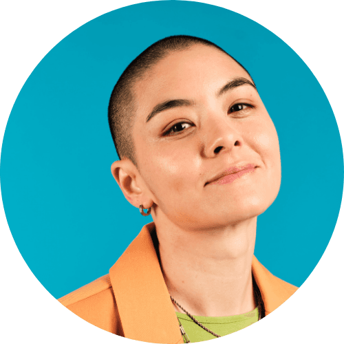 Portrait of a smiling woman with a shaved head, wearing an orange blazer and green top, with a blue background.