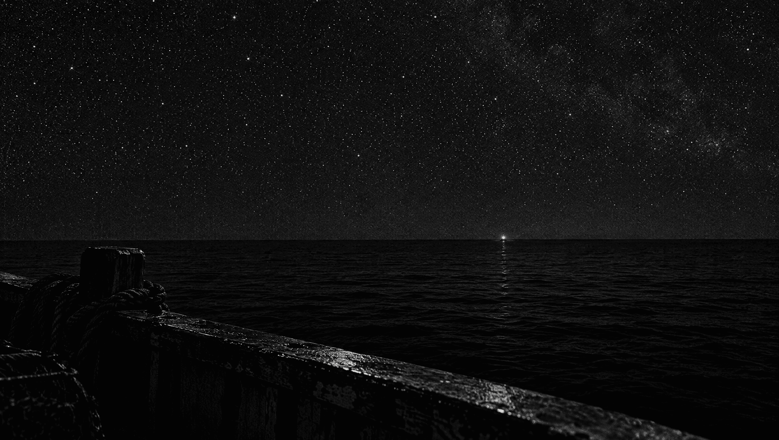 Night at sea under a sky full of stars, a single small light low on the far horizon