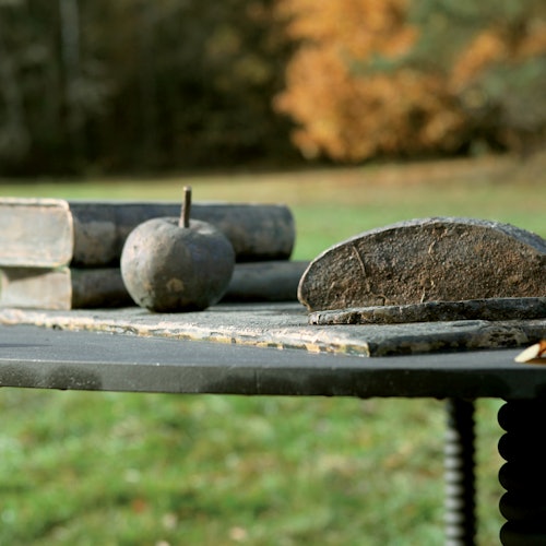 Outdoor table with old books, a round stone, and a broken grindstone, set against an autumn background with trees and grass.