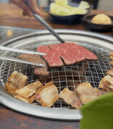 A thick cut of steak with prominent grill marks sizzles over hot coals and flames on a barbecue, with blurred figures of people and drinks in the background, suggesting an outdoor gathering.