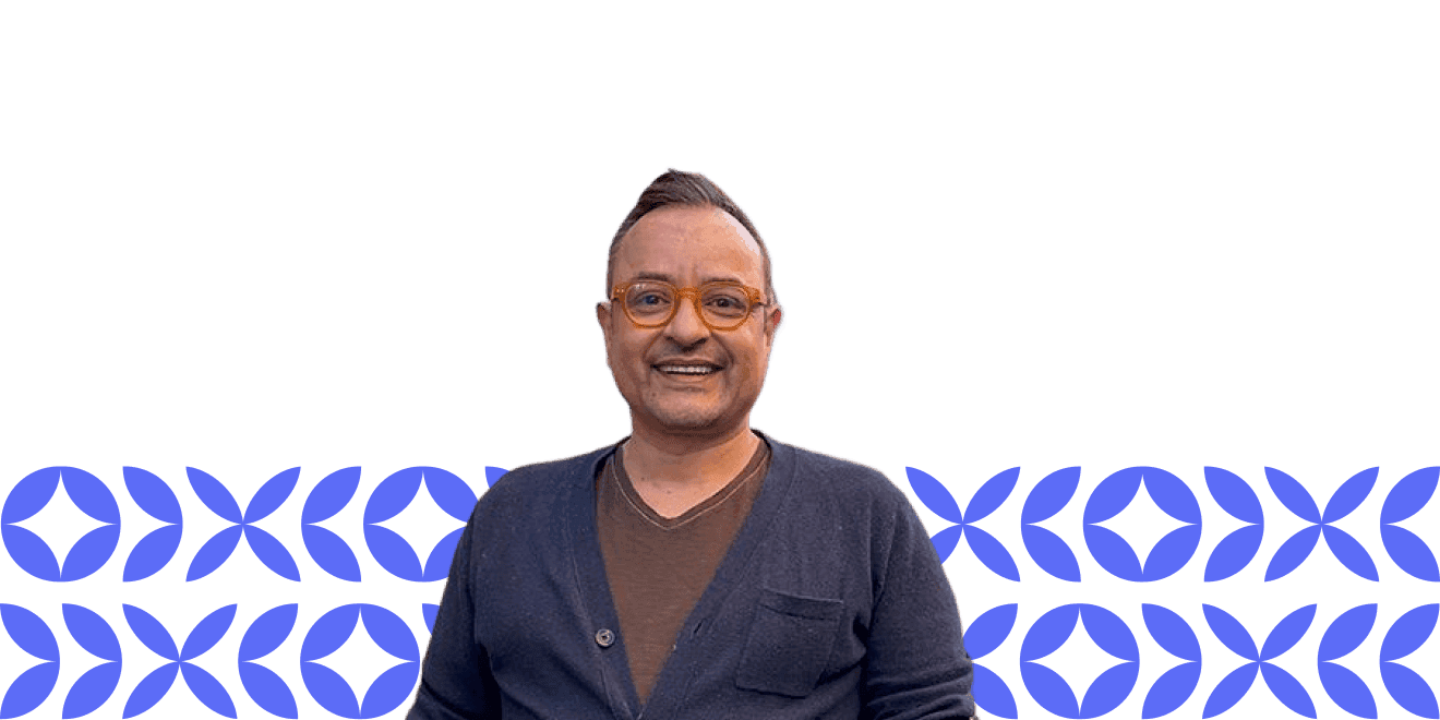 Um homem usando óculos laranja e um cardigã azul-marinho sorri contra um fundo de padrões geométricos azuis.