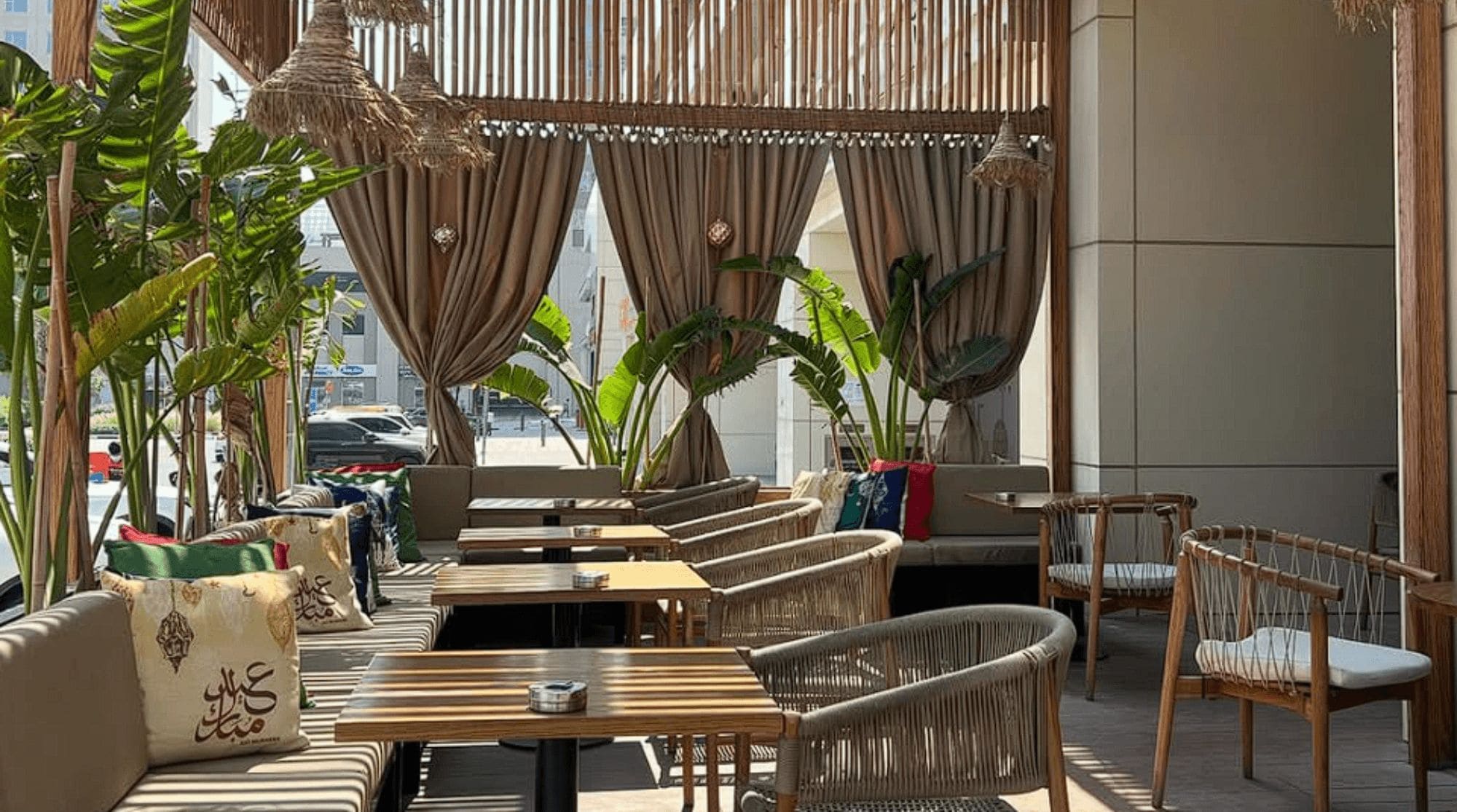 A modern breakfast place in Dubai with a full outdoor seating, brown curtains, cushioned seats, wooden tabletops, and greenery.