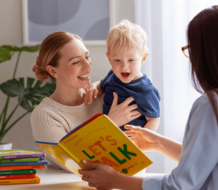 Speech therapist reading a colorful book titled 'Let’s Talk' with a smiling toddler and mother during a language therapy session.