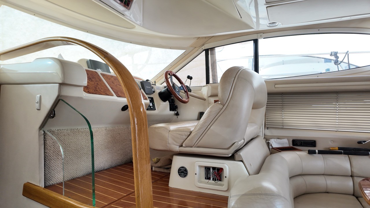 Sealine F37 interior lower helm station and saloon — cream leather captain's chair, wood-trimmed steering wheel, Raymarine navigation electronics, teak sole flooring, and panoramic wraparound windows with venetian blinds