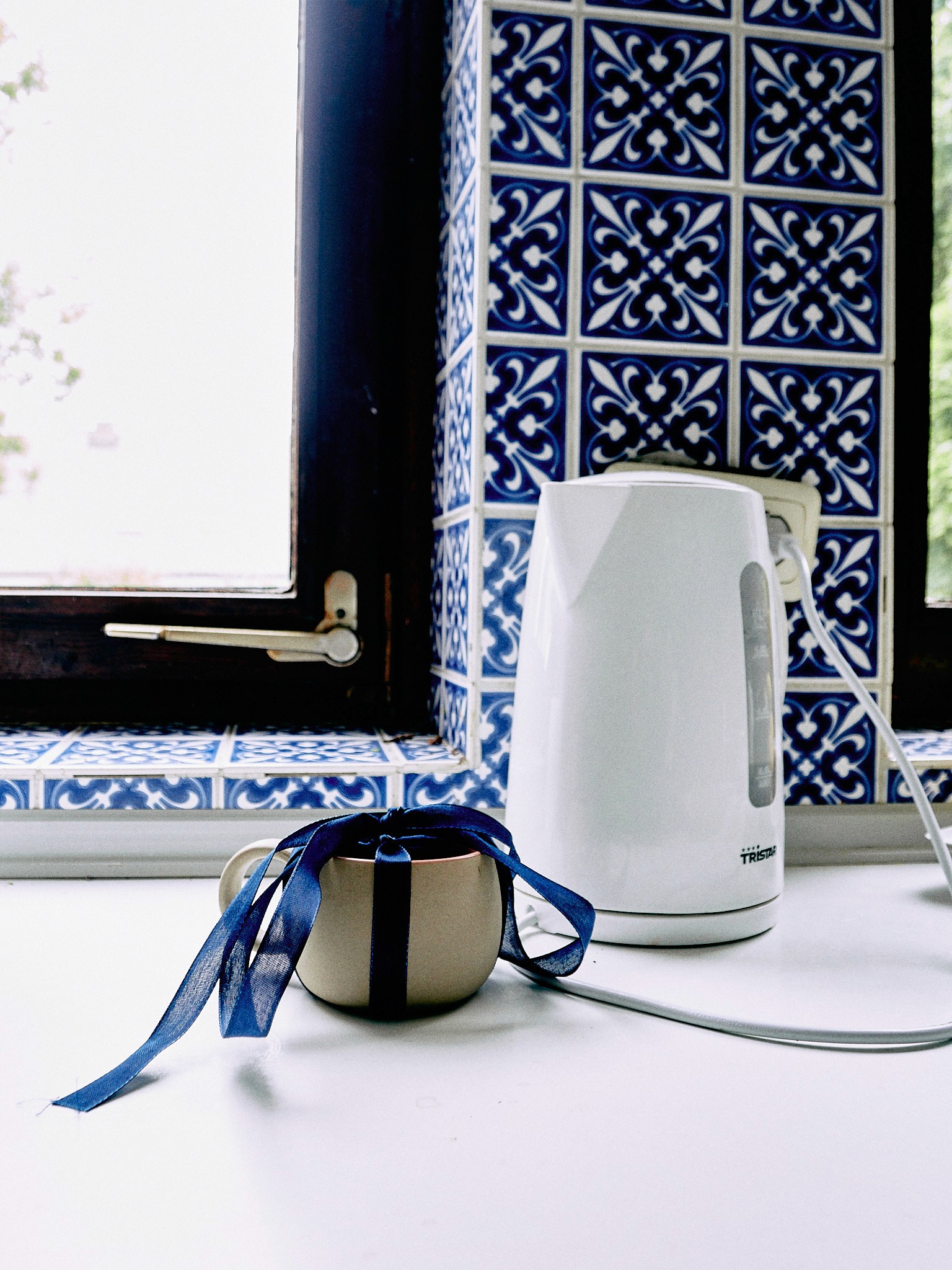 Beige Tasse mit blauem Band auf weißer Fensterbank neben weißem Wasserkocher vor blau-weißen Fliesen – Alltagsfotografie von Ovid Dernbach. 