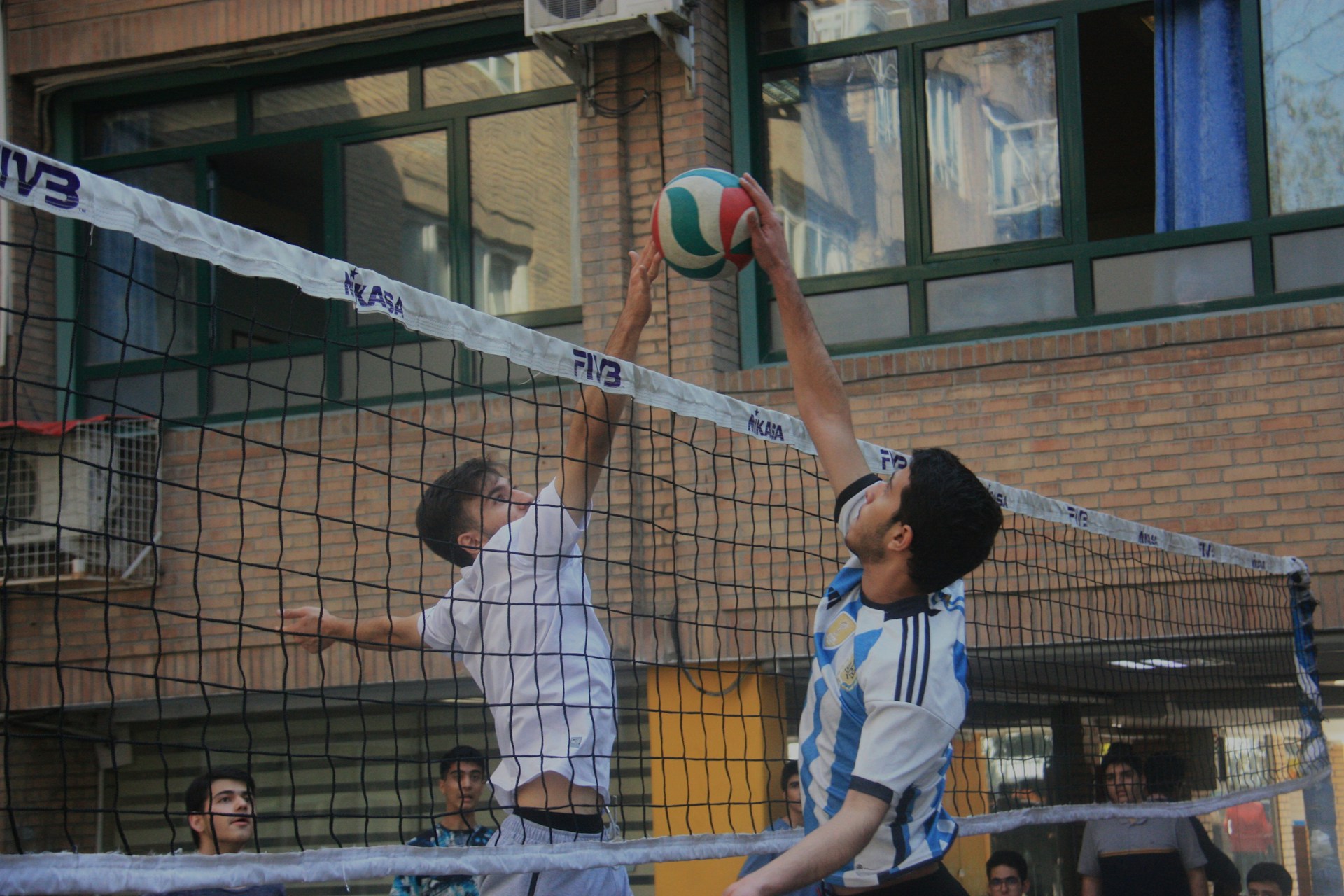 Two volleyball players jumping at the net to contest the ball during an outdoor game.
