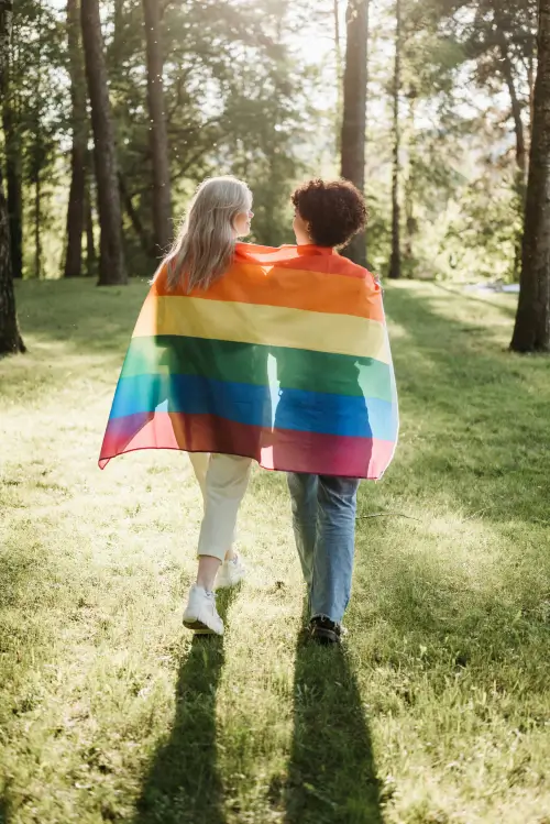 Back view shot of women walking in a sunlit park with a rainbow flag wrapped around their shoulders.