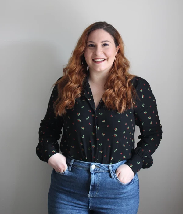 A person standing against a plain, light‑coloured wall in a relaxed, confident pose, with both hands in the pockets of high‑waisted blue jeans. They are wearing a black long‑sleeved blouse patterned with small, colourful motifs. Their long, wavy auburn hair falls over their shoulders. The image is softly lit and framed from mid‑thigh upwards, giving a clean, editorial‑style portrait suitable for a profile or team page.