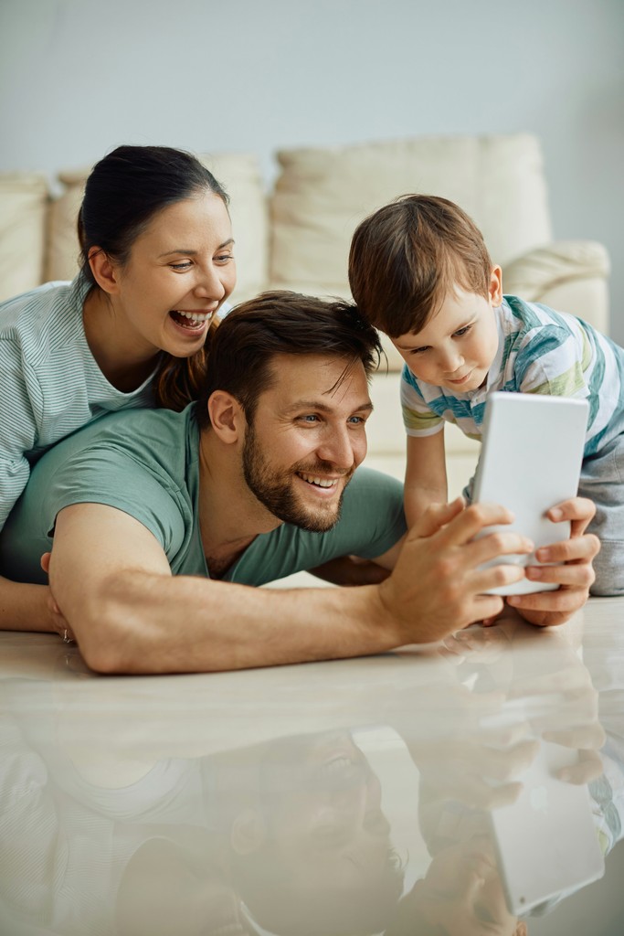 The image features a joyful family of three—parents and their young son—lying on the floor and smiling as they look at a tablet together. The relaxed home setting, with a beige couch in the background, adds to the warmth and happiness of the moment, capturing a sense of togetherness and digital bonding.