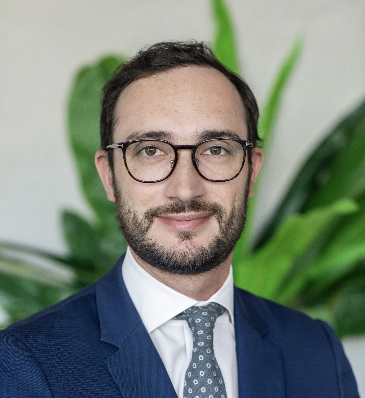 A smiling man with curly hair is sitting at a desk, looking at the camera, with a laptop and a lamp in the background.