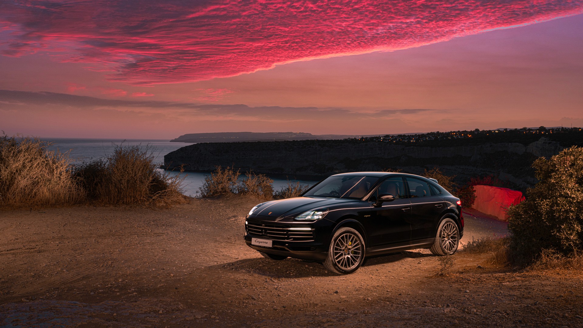 Black Porsche Cayenne parked on a dirt clifftop above the sea at dusk, a vivid pink-and-red cloudbank overhead, with coastline and distant town lights along the bay behind.
