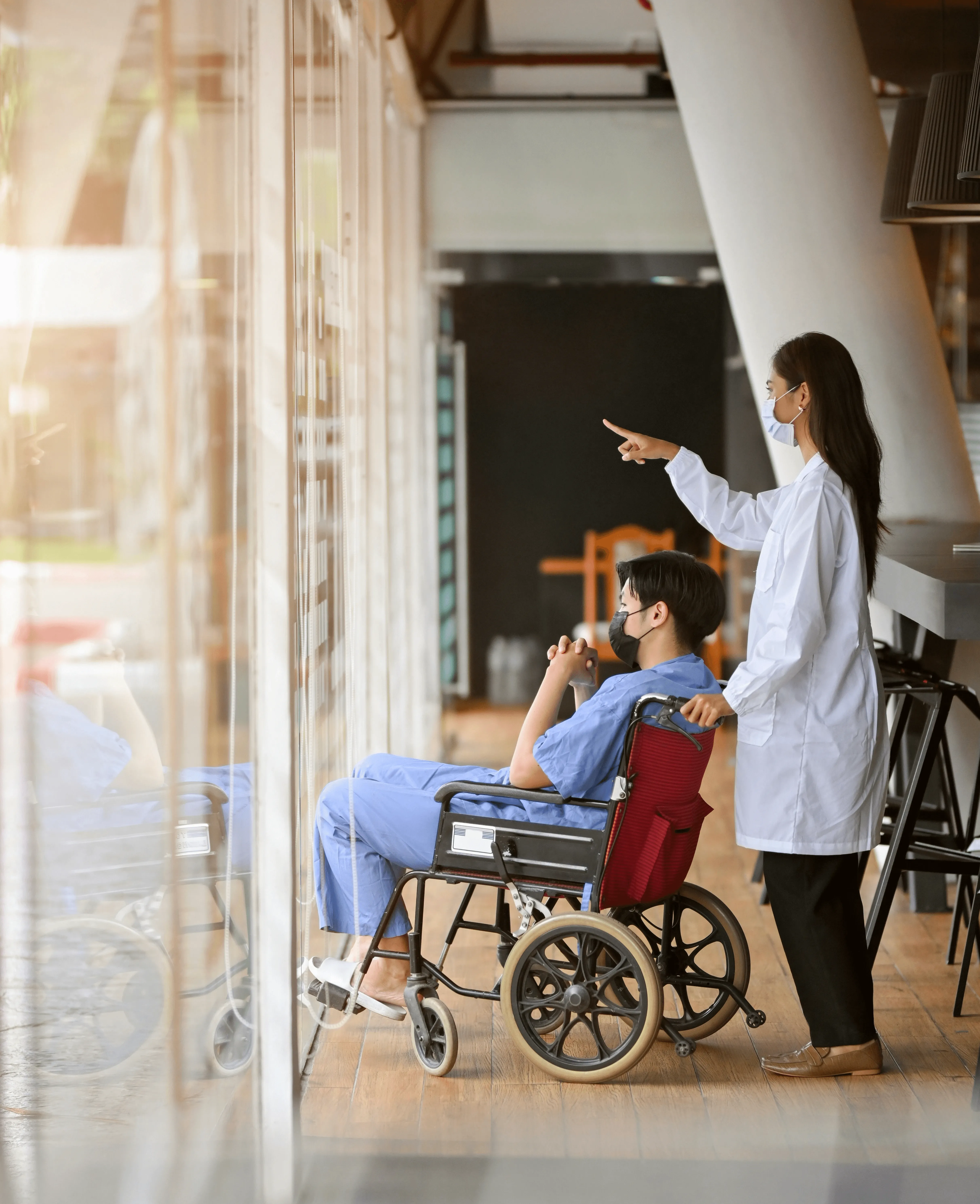 A healthcare worker points to a display while assisting a patient in a wheelchair in a hospital setting.