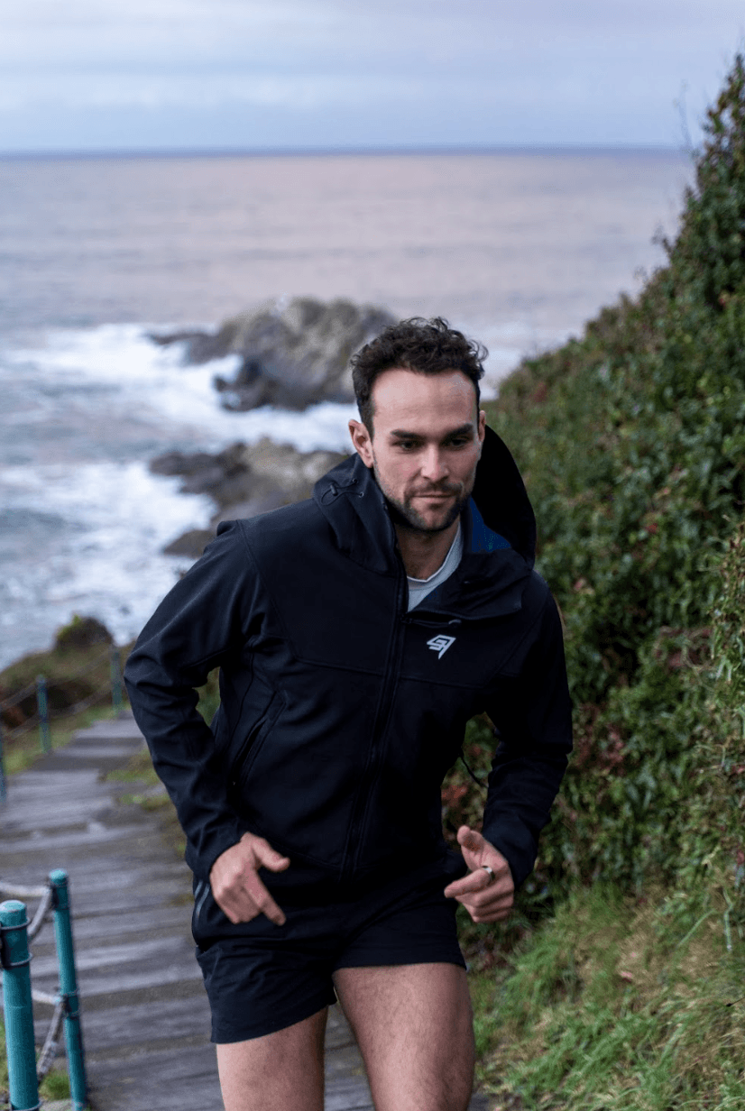 Man wearing olive green and orange sneakers with tall socks, standing on an urban street with a modern fence and skyscraper in the distance.