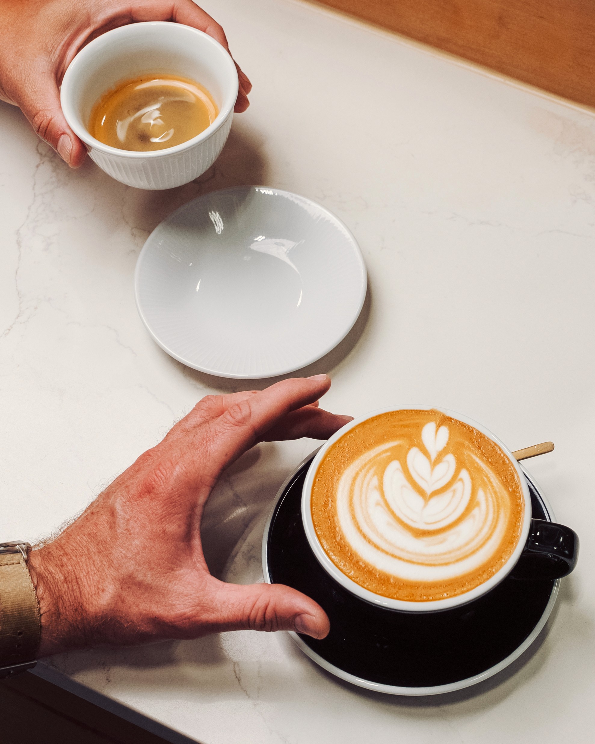 A hand reaches for a latte with intricate foam art, while another hand holds a smaller cup on a table.