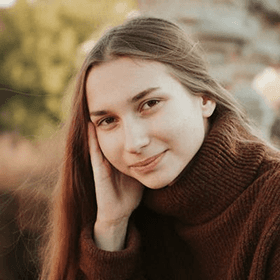 A young woman in a cozy sweater smiles gently while resting her hand on her cheek, surrounded by autumn foliage.
