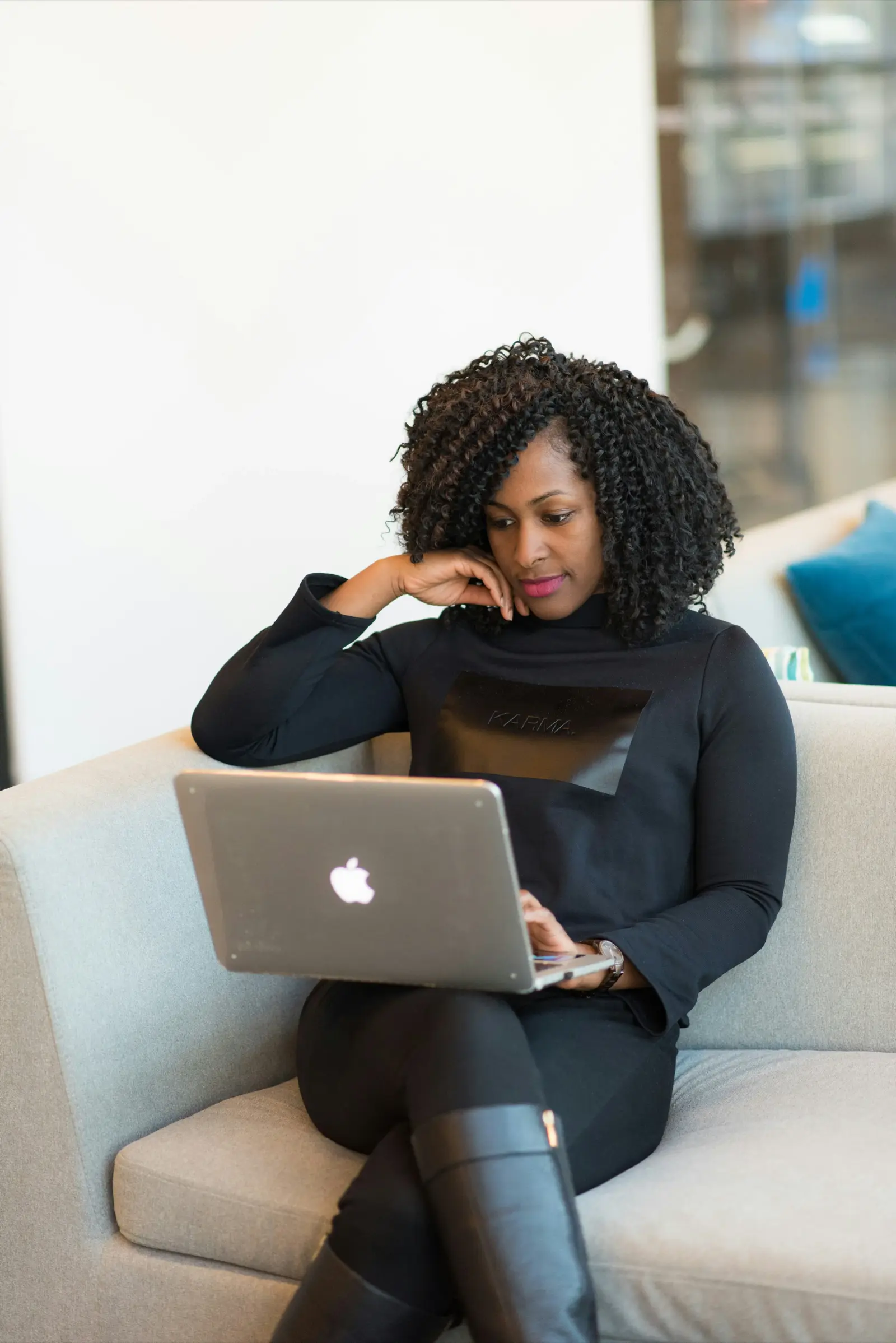 A person with curly hair sits on a couch, focused on a laptop while resting their chin on their hand.