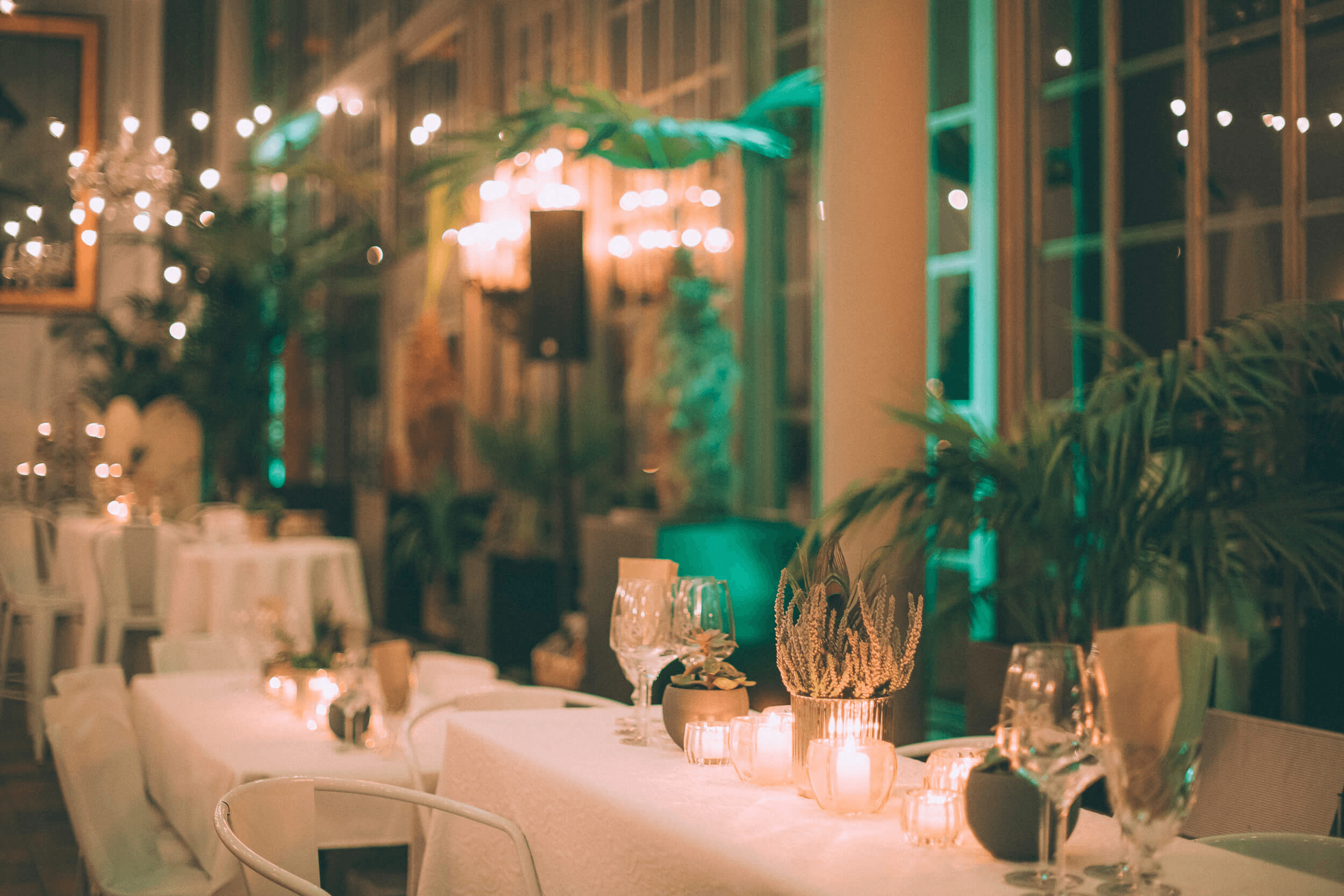 Outdoor restaurant terrace overlooking a tranquil lake at sunset. White tables and chairs are set with glassware. Sheer curtains add elegance. Calm ambiance.