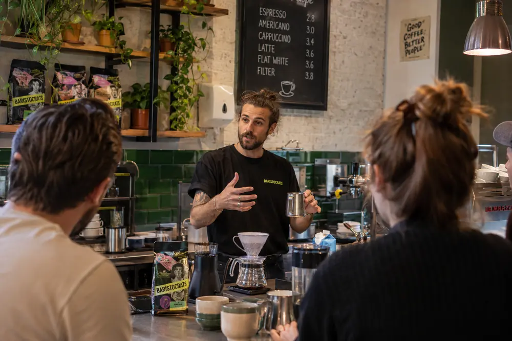 Baristocrats trainer explains pourover technique to a group at a coffee bar.