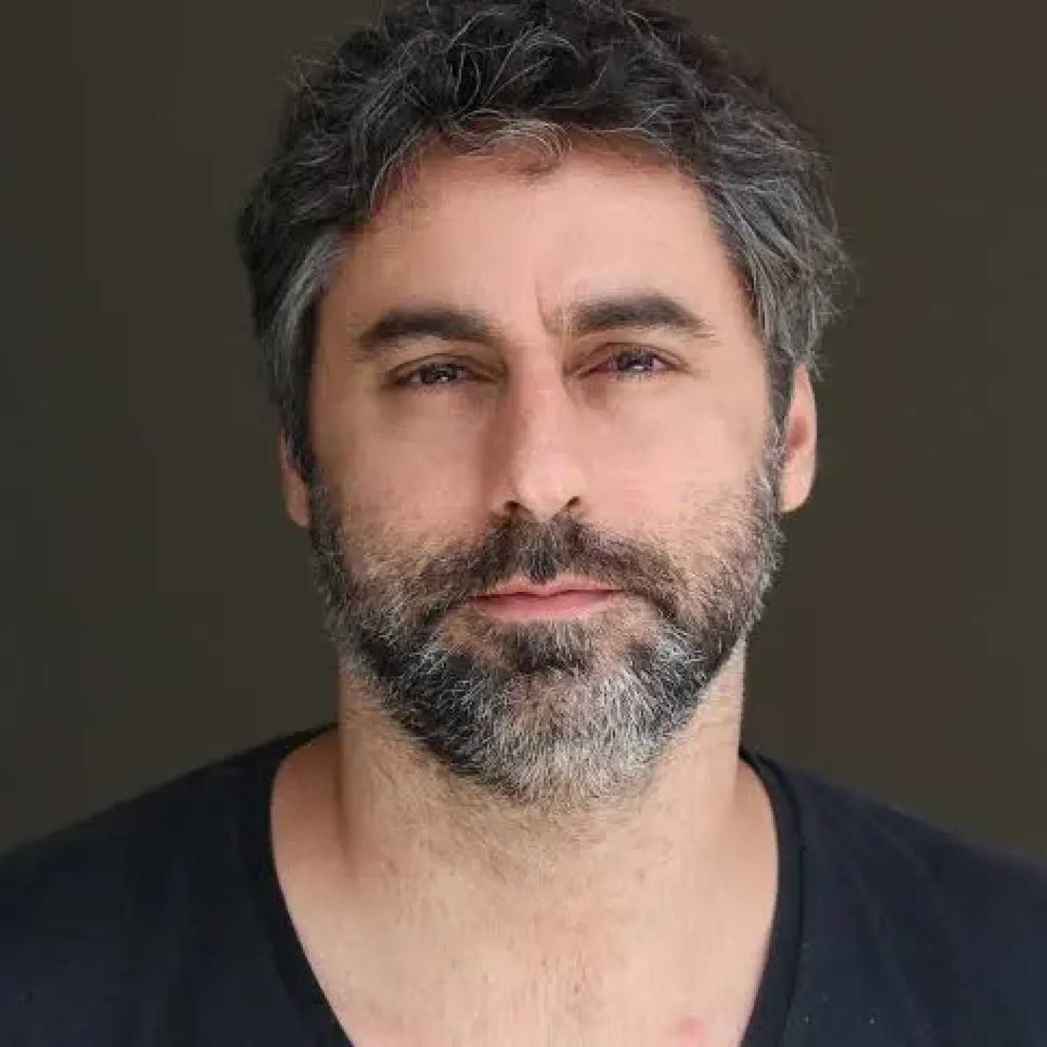 A young person with curly hair smiles at the camera while sitting in front of a laptop against a dark background.