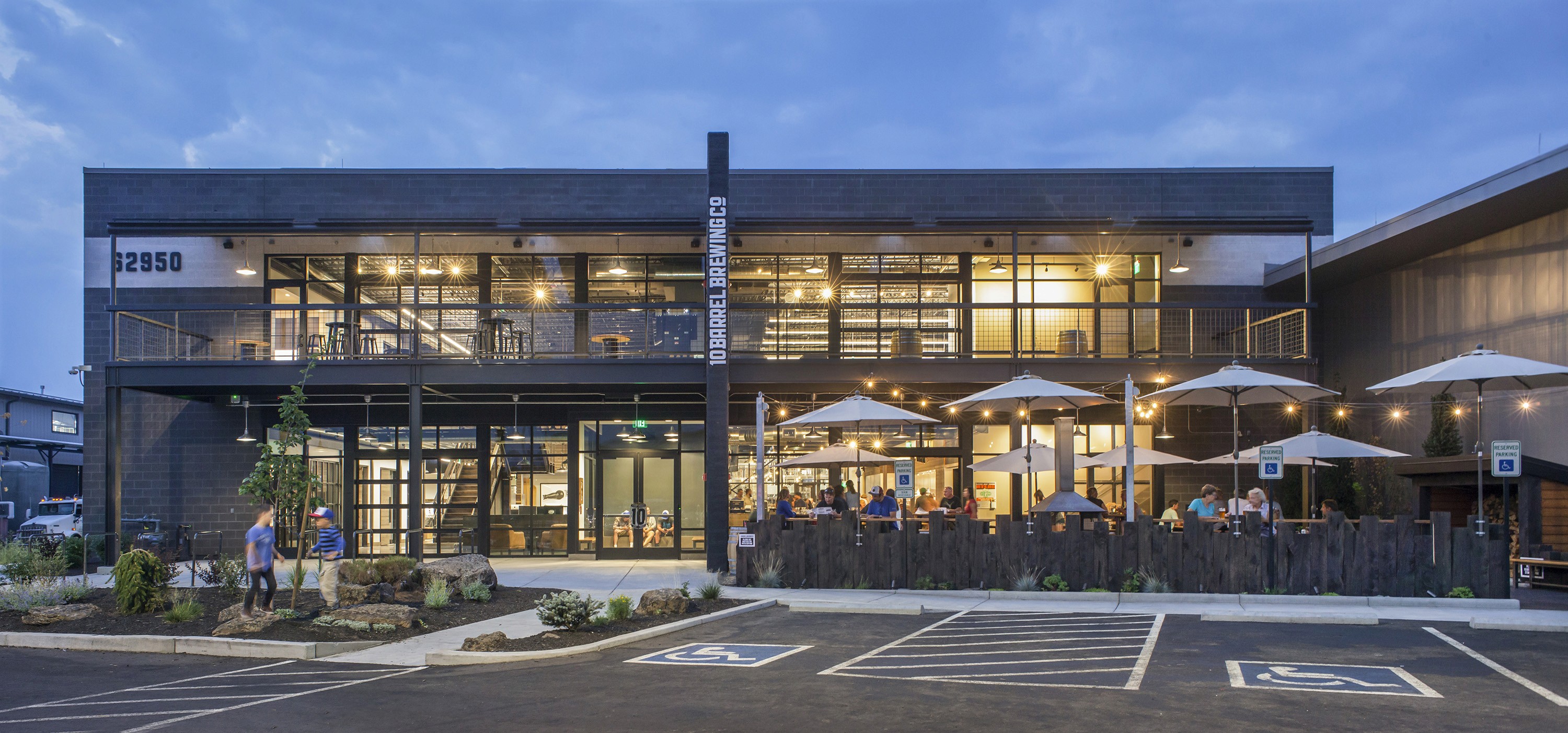 Two-story glass and dark-metal brewery headquarters at dusk, with outdoor seating along the front