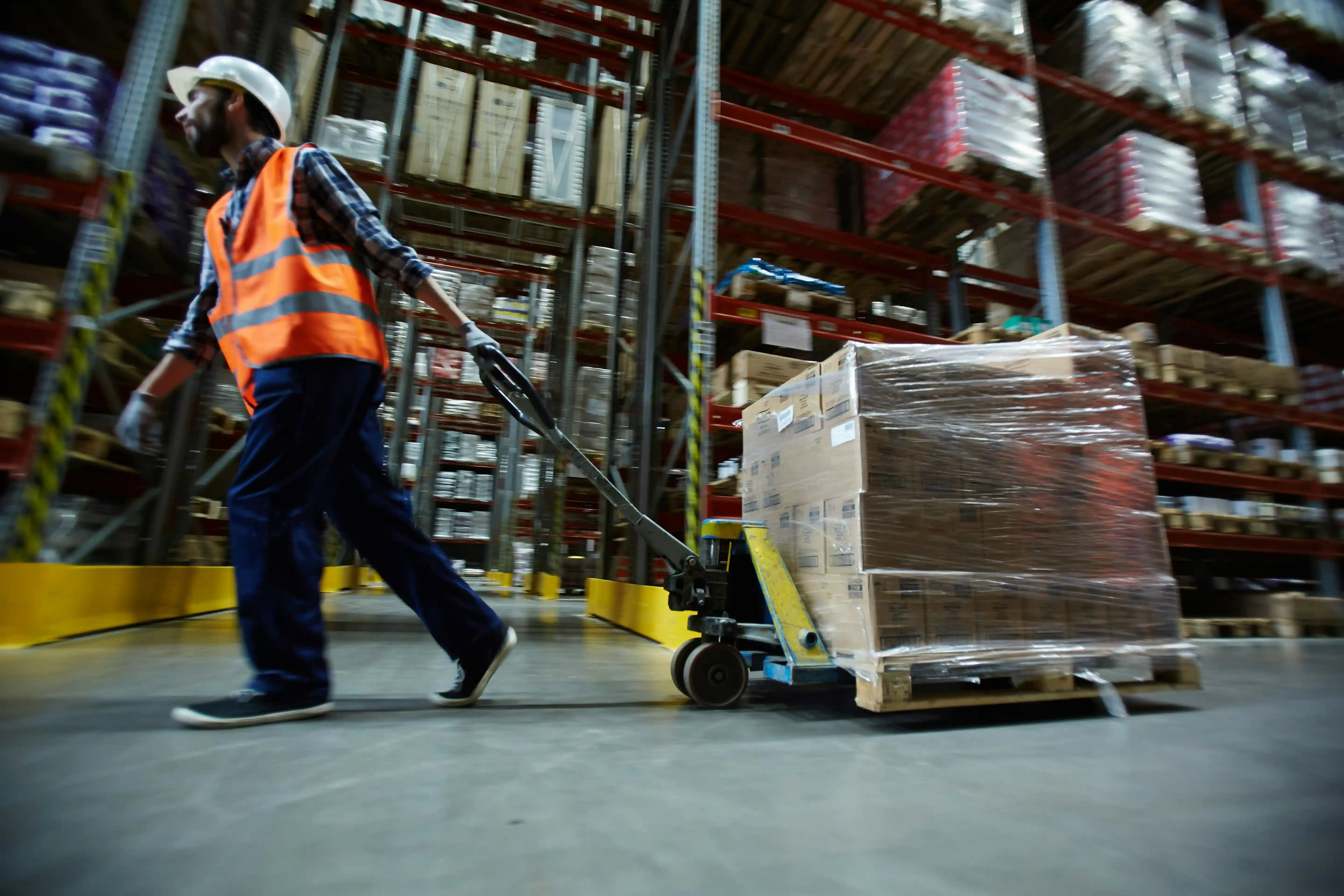 a warehouse worker in a bight orange high-vis jacket drags a pallet trolley through a busy 3PL warehouse