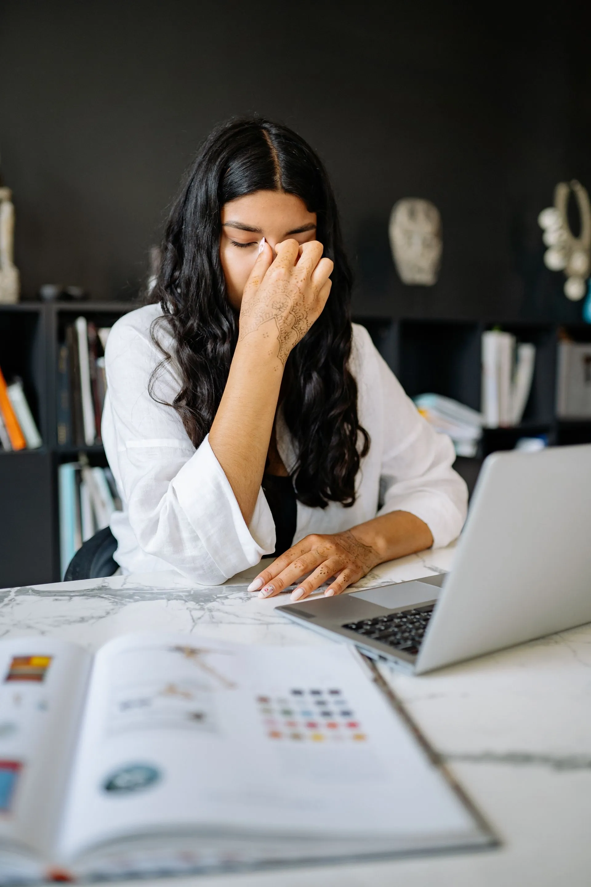 A woman with long dark hair sits at a desk in front of a laptop, rubbing her eyes in frustration, surrounded by books and a dark, stylish office background.