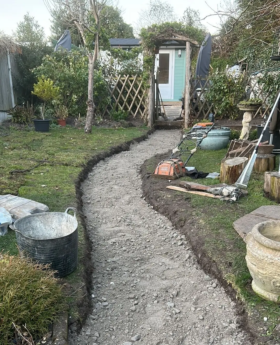 A winding gravel path leading through a lush garden with plants and garden structures on either side.