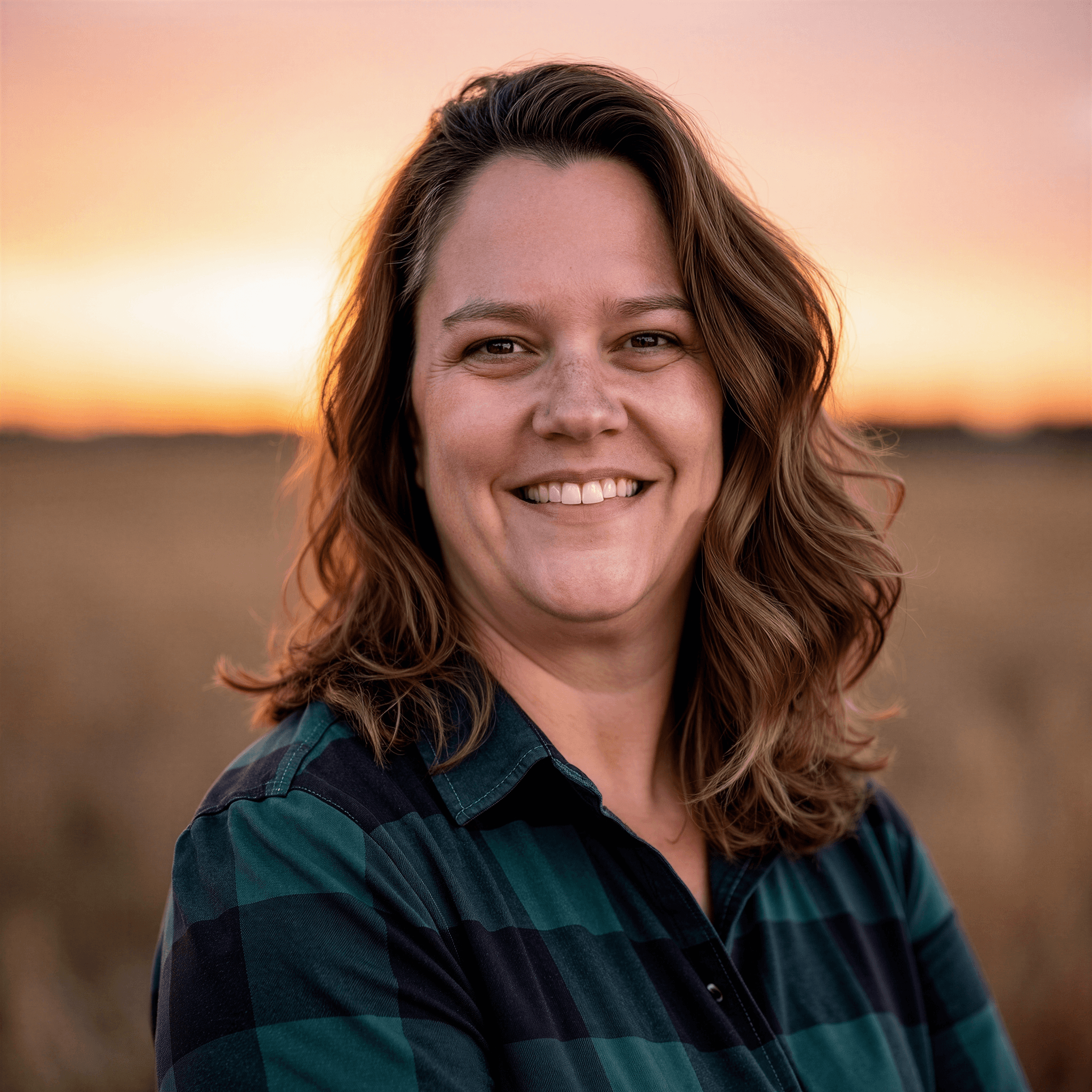 Smiling woman in plaid shirt outdoors at sunset, portrait highlighting warmth, resilience, and personal connection.