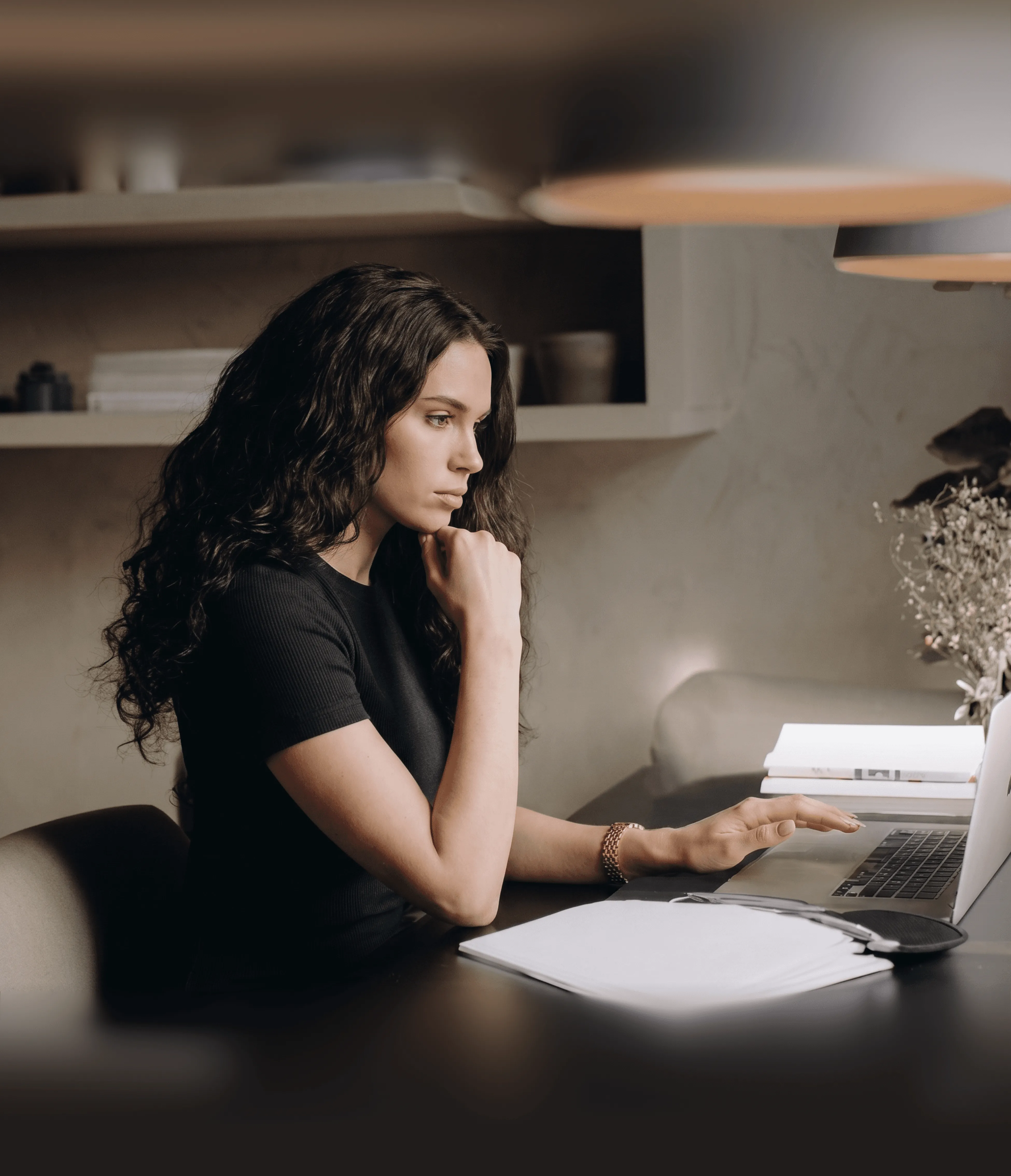 A person with long, curly brown hair sits thoughtfully at a desk, focusing on a laptop, with notebooks and a planner nearby, in a softly lit, modern home office setting.