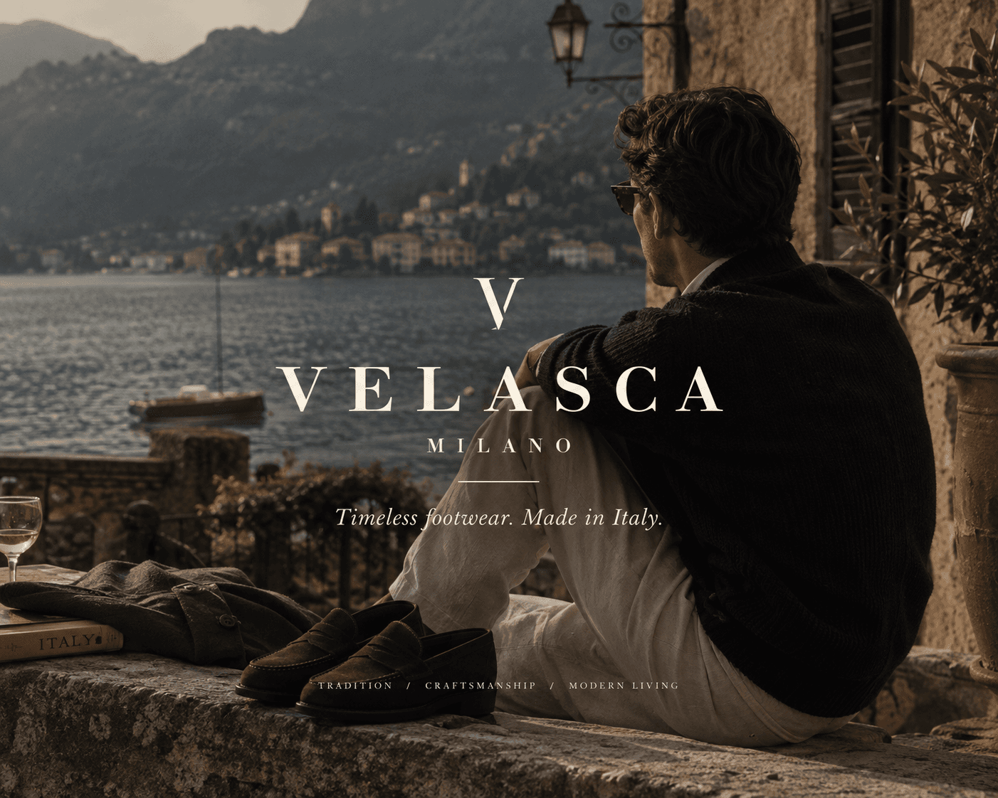 A back profile view of a man wearing a dark navy cardigan and white trousers sitting on a stone terrace wall overlooking a picturesque Italian lake framed by mountains. Beside him on the ledge sit dark brown suede loafers, an olive jacket, a wine glass, and a book titled "ITALY". Across the center, white brand text displays "V VELASCA MILANO", the tagline "Timeless footwear. Made in Italy.", and "TRADITION / CRAFTSMANSHIP / MODERN LIVING".