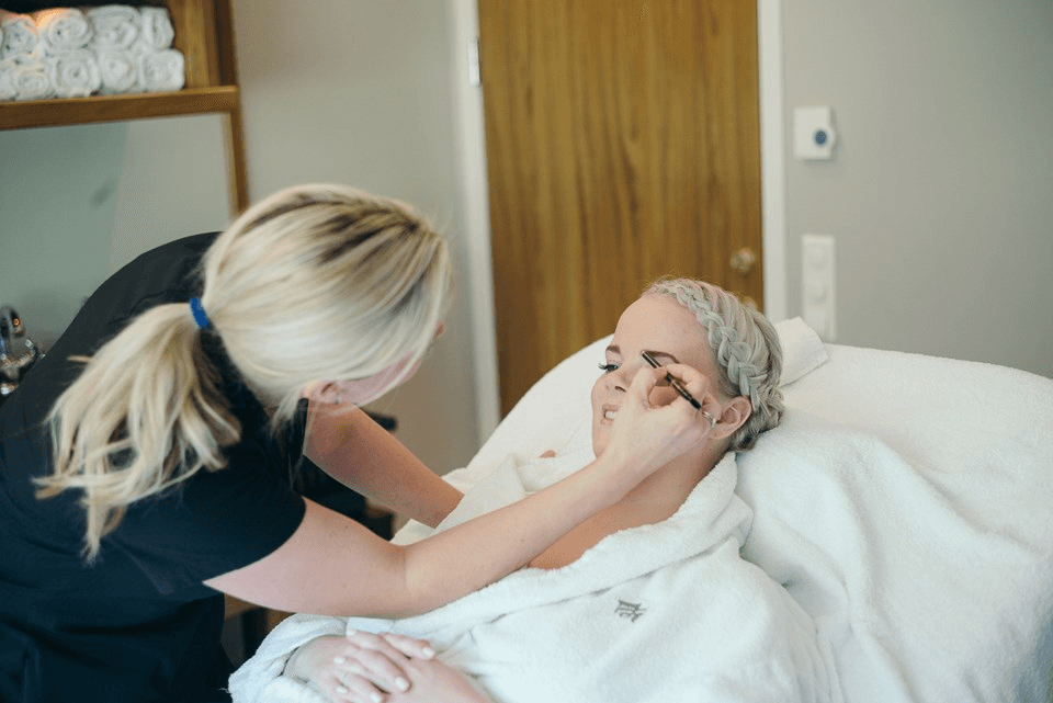 A woman receives a professional makeup application while lying in a spa treatment room, wrapped in a plush white robe, with neatly folded towels in the background.